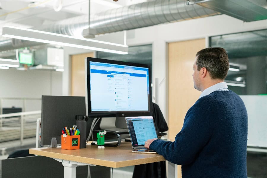 A man standing at a height-adjustable desk, working on a laptop and viewing LinkedIn on a large desktop monitor in a modern office setting.