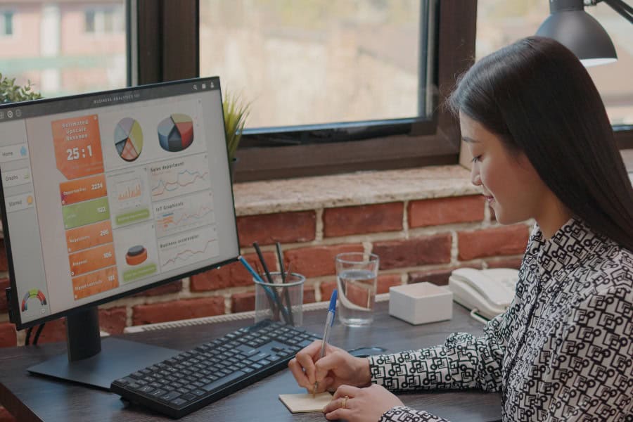 Woman taking notes while analysing data charts on a computer screen in a modern office.