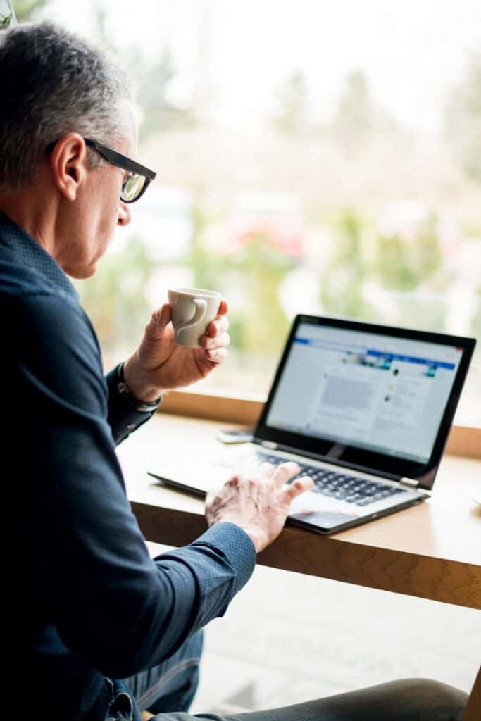 Man in glasses working on a laptop with a coffee cup in hand, seated by a bright window.