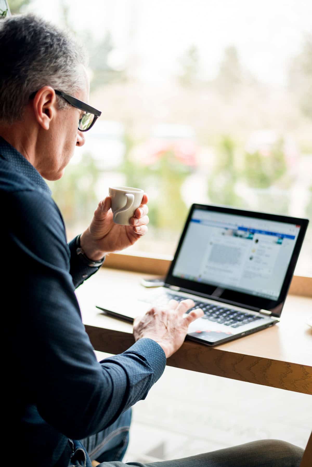 Man in glasses working on a laptop with a coffee cup in hand, seated by a bright window.